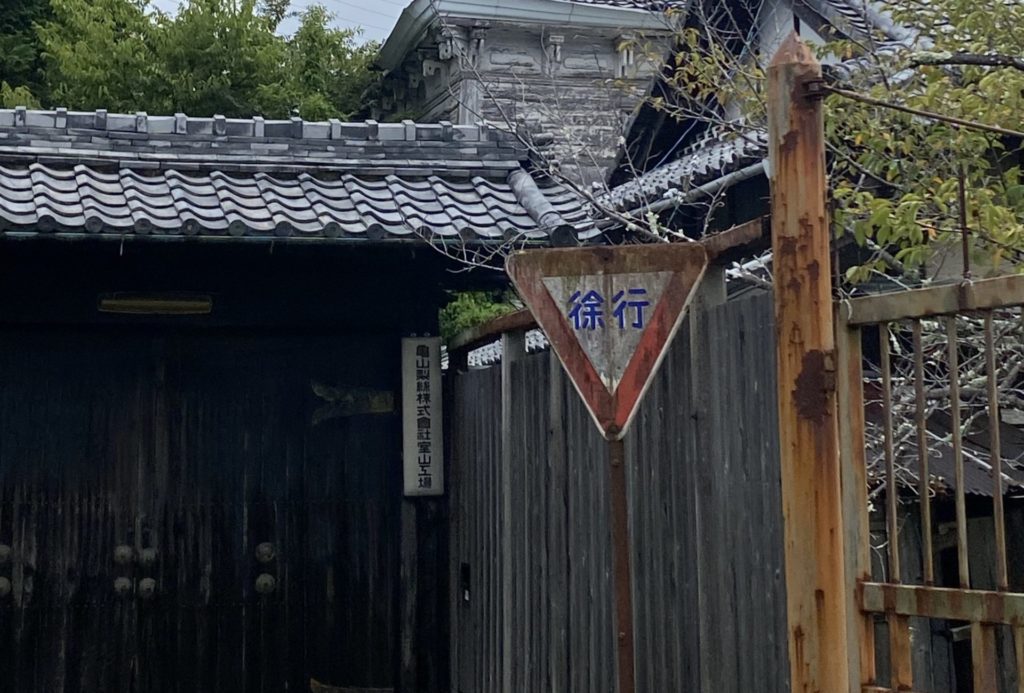 徐行と交通標識の後に瓦の乗った門があり、「亀山製糸株式会社室山工場の看板がかかっている