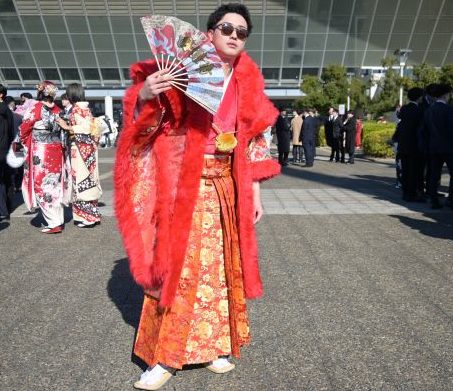 赤いファーの付いた羽織で赤い袴の男性が四日市と書いた扇子を持ち、サングラスをして立っている